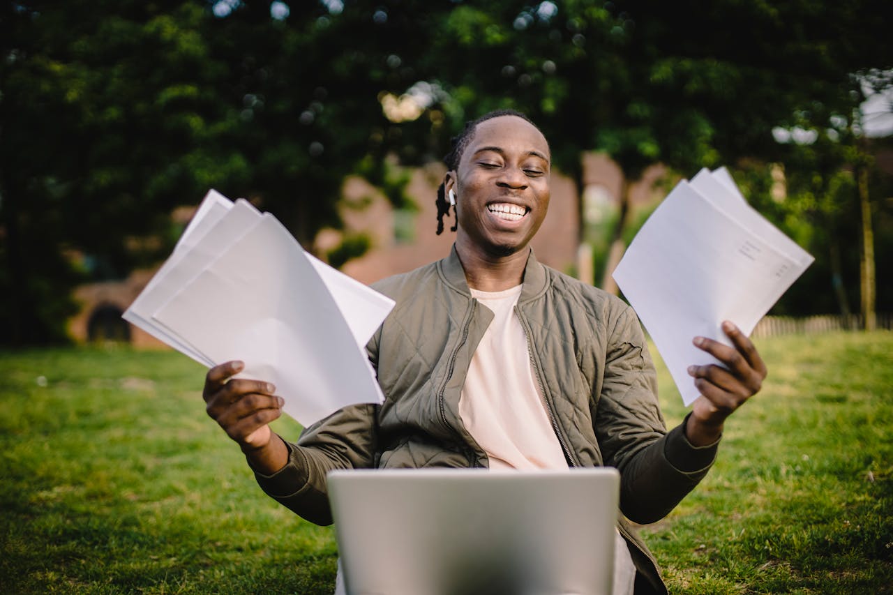 Happy young man holding papers with a laptop outdoors, showcasing academic achievement and joy.